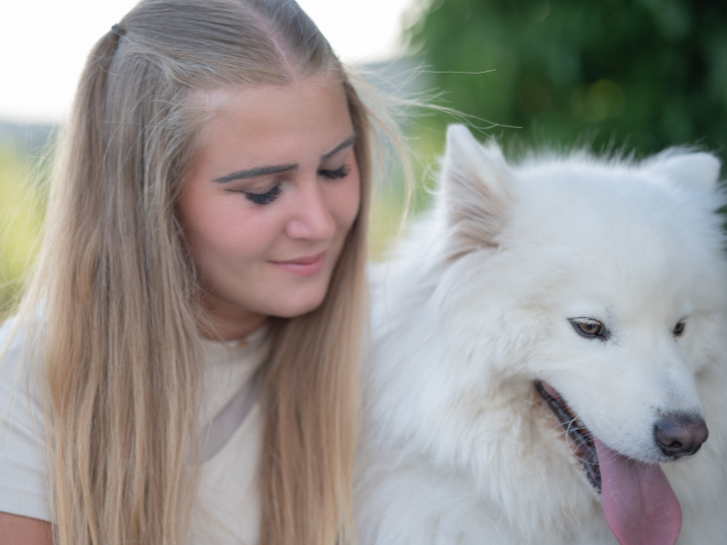 Eine blonde junge Frau kuschelt mit ihrem weißen Samoyede und hat dabei genießerisch die Augen geschlossen.